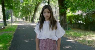 Düşünceli bir kız, sakin, ağaçlarla kaplı bir parkta tek başına dikiliyor, kameraya bakıyor, günlük beyaz bir bluz ve desenli bir etek giyiyor.