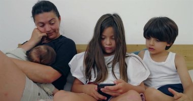 Bir anne, büyük çocukları telefona odaklanırken bebeğini emziriyor, yatakta samimi bir aile anı yaratıyor, evde bir birliktelik ve rahatlama hissi yaratıyor.