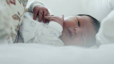 Yeni doğmuş bebek yatakta ağlıyor, rahatsız edici bir şekilde elini kaldırıyor. Bebeğin yüzünde sıkıntı var, ve jestler tedirginliği gösteriyor.