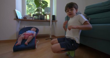 İki küçük çocuk, fedaide yatan bir bebek ve yakınlarda oturan büyük bir kardeş, rahat bir oturma odasında, kardeş bağı ve aile hayatı sergiliyorlar.