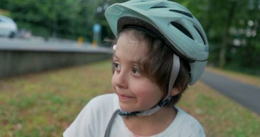 Kaykaylı bir kaykayın üzerinde oturan, düşünceli görünen, başlık takan, bulutlu bir günde ağaçlar ve yeşilliklerle çevrili bir çocuk.