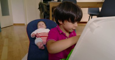 Büyük kardeş oyuncakla oynarken küçük kardeş bir fedainin içinde dinleniyor, aile hayatının bir anını yakalıyor ve rahat bir ev ortamında kardeş etkileşimi kuruyor.