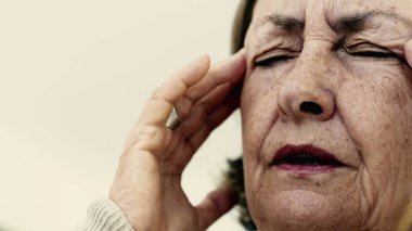 Yaşlı bir kadın şakaklarına sıkıca masaj yapıyor. Acıyı veya stresi azaltmaya odaklanıyor. Derin kırışıklıklar, yoğun yüz ifadesi, görülebilir gerilim şiddetli baş ağrısı iletir.