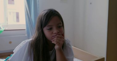 Genç bir kız, parlak bir odada renkli eşyalarla çevrili, dalgın bir ifadeyle yatağında oturuyor, bir anlık tefekkür ve içgözlem anı yakalıyor.