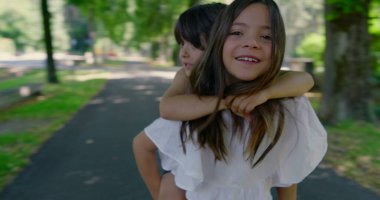 Kız kardeşi, güneşli bir günde erkek kardeşini sırtında taşıyor. Hem gülüyor, hem de bahçenin tadını çıkarıyor. Etrafı yeşillik ve gölgeli yürüyüş yolu ile çevrili.