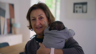 Büyükanne yeni doğmuş bir bebeği omzunda tutuyor, kameraya sıcak bir şekilde gülümsüyor, sıcak bir ev ortamında aile sevgisini ve bağlantısını gösteriyor.