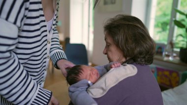 Anne, yeni doğan bebeğini büyükannesinden almak için uzanır, sıcak bir aile bağı ve bakım anını yakalar.