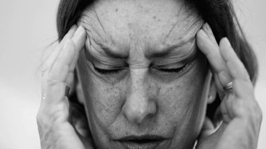 Yaşlı bir kadın parmaklarını şakaklarına bastırıyor, gözleri kapalı, şiddetli baş ağrısı ve stres belirtileri gösteriyor. Derin kırışıklıklar ve sıkı yüz ifadesi rahatsızlığı gösteriyor.