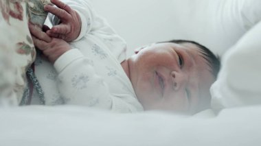 Yeni doğmuş bebek yatakta ağlıyor, rahatsız edici bir şekilde elini kaldırıyor. Bebeğin yüzünde sıkıntı var, ve jestler tedirginliği gösteriyor.