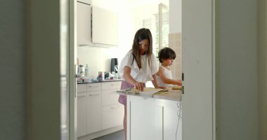 Mutfakta kardeşler yemek hazırlıyor, kız ve oğlan yemek pişirmeye odaklanıyor, aşçılık faaliyetleri, aile ilişkileri, arka planda anne, ışık dolu mutfak.