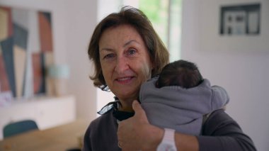 Büyükanne yeni doğmuş bir bebeği omzunda tutuyor, kameraya sıcak bir şekilde gülümsüyor, sıcak bir ev ortamında aile sevgisini ve bağlantısını gösteriyor.