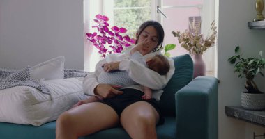 Anne bebeğini kucağında, bebek yüzüne dokunurken yavaşça kanepede dinleniyor. Huzurlu bir ev ortamında samimi bir yakınlık ve bağ anı.