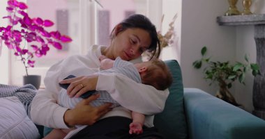 Anne bebeğini kanepede tutuyor, yüzü hem yorgunluk hem de sakinlik gösteriyor, anne olmanın günlük gerçekliğini yansıtıyor, rahat ve huzurlu bir yuvada.
