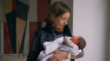 Büyükanne sevgiyle kundaklanmış yeni doğmuş bir bebeği kollarında tutuyor. Büyükannem, bebekle ilgilenirken şefkatle aşağı bakar. Bir anlık bağ ve özen gösterir.