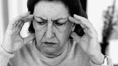 Şiddetli baş ağrısı ile mücadele eden yaşlı bir kadın, gözle görülür konsantrasyonda şakaklara masaj yapıyor. Yüz hatları ve yüz ifadeleri yoğun acıyı, zihinsel gerginliği, duygusal sıkıntıyı yansıtır.