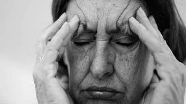 Yaşlı bir kadın parmaklarını şakaklarına bastırıyor, gözleri kapalı, şiddetli baş ağrısı ve stres belirtileri gösteriyor. Derin kırışıklıklar ve sıkı yüz ifadesi rahatsızlığı gösteriyor.