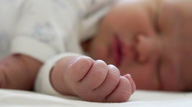 Bebeğin yumuşaklığını ve hayatın ilk anlarını vurgulayan, uyuyan bebeğin bulanık arkaplanlı arka planına sahip, yeni doğmuş bebeklerin minik yumruğuna yakın çekim