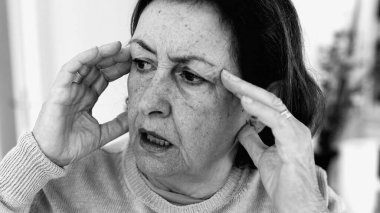 Şiddetli baş ağrısı ile mücadele eden yaşlı bir kadın, gözle görülür konsantrasyonda şakaklara masaj yapıyor. Yüz hatları ve yüz ifadeleri yoğun acıyı, zihinsel gerginliği, duygusal sıkıntıyı yansıtır.