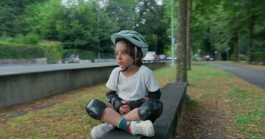 Koruyucu teçhizatlı bir çocuk yol kenarında kaykayın üzerinde oturuyor, kaskını takıyor, geçen arabalara bakıyor, yeşil ve ağaçlarla çevrili, bulutlu bir günde.