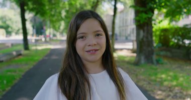 Parkta ağaçlarla kaplı bir yolda duran genç kız, kameraya tarafsız bir ifadeyle bakıyor, yeşil ve güneş ışığıyla çevrili, sakin bir açık hava anını yakalıyor.