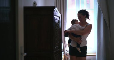 Anne bebeğini hafifçe aydınlatılmış bir odada kucağında tutuyor, bebeği huzurlu, şefkatli bir şekilde kucaklıyor ve onunla bağlantı kuruyor, sevgisini ve sakinliğini gösteriyor.