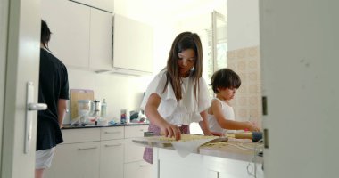 Mutfakta kardeşler yemek hazırlıyor, kız ve oğlan yemek pişirme işine odaklanıyor, aşçılık faaliyetleri, aile içi ilişkiler, arka planda anne, ışık dolu mutfak-SD 480p