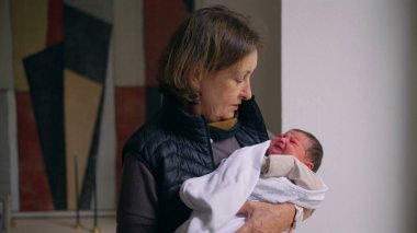 Büyükanne sevgiyle kundaklanmış yeni doğmuş bir bebeği kollarında tutuyor. Büyükannem, bebekle ilgilenirken şefkatle aşağı bakar. Bir anlık bağ ve özen gösterir.