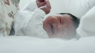 Yeni doğmuş bebek yatakta ağlıyor, rahatsız edici bir şekilde elini kaldırıyor. Bebeğin yüzünde sıkıntı var, ve jestler tedirginliği gösteriyor.