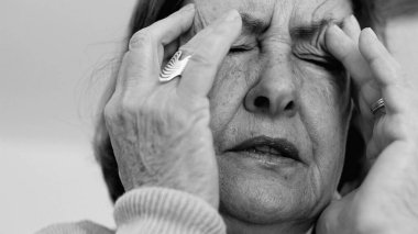 Yaşlı kadın yakın plan, gözleri kapalı, iki eli şakaklarına bastırıyor, derin kırışıklıklar ve acı dolu ifade, şiddetli stres ya da baş ağrısı