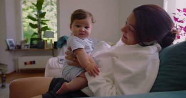 Anne, güneşli bir oturma odasında bebeğine gülümserken anne ve çocuk arasında sıcak ve sevgi dolu bir bağ kuruyor.
