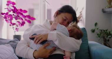 Anne bebeğini kanepede tutuyor, yüzü hem yorgunluk hem de sakinlik gösteriyor, anne olmanın günlük gerçekliğini yansıtıyor, rahat ve huzurlu bir yuvada.