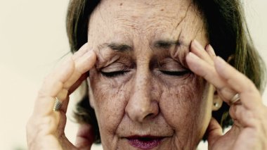 Yaşlı bir kadın şakaklarına sıkıca masaj yapıyor. Acıyı veya stresi azaltmaya odaklanıyor. Derin kırışıklıklar, yoğun yüz ifadesi, görülebilir gerilim şiddetli baş ağrısı iletir.