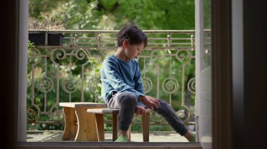 Genç çocuk bir balkonda ahşap bir taburede oturuyor, düşünceli bir şekilde aşağı bakıyor, çevrende yeşil bir manzara, ağaçların arasından süzülen doğal ışık.