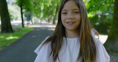 Uzun saçlı bir kız parkta kameraya bakarken geri geri yürüyor, beyaz bir bluz giyiyor, arka planda küçük bir çocuk oynuyor, ağaçlar ve yeşilliklerle çevrili.