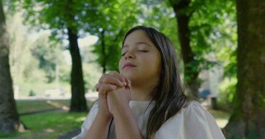 Parkta duran uzun saçlı genç bir kız, gözleri kapalı ve elleri dua ederken sımsıkı sarılmış, yemyeşil ve ağaçlarla çevrili, çocukluk maneviyatını ve barışçıl tefekküre önem veren