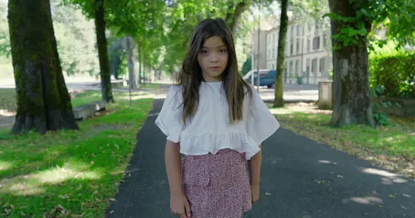 Mutlu kız, yeşil bir parkta, ağaçlarla kaplı bir yolda duruyor, kameraya sıcak bir şekilde gülümsüyor, beyaz bir bluz ve desenli bir etek giyiyor, güzel bir günün tadını çıkarıyor.