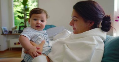 Bebeğin annesinin kollarındayken kameraya baktığı yakın plan, bebeğin parlak, ev ortamındaki meraklı ve nişanlı ifadesi..