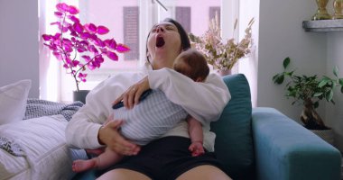 Anne, bebeğini koltukta emzirirken esniyor. Rahat bir ev ortamında, ebeveynlikte samimi bir yorgunluk ve sevgi anı yakalıyor.