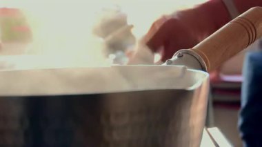 Çekiçle dövülmüş metal bir tencereden yükselen buhar, ahşap saplı, mutfakta pişirme işlemi, kaynayan sıvı, yemek hazırlama, sıcak tencere, aşçılık sahnesi, ev yemeği.