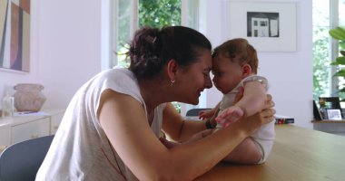 Anne sevecen bir şekilde bebeğini kucağında tutuyor, alnına dokunuyor ve doğal ışıklandırılmış bir ev ortamında ahşap bir masada duygusal bir an yaşıyor.
