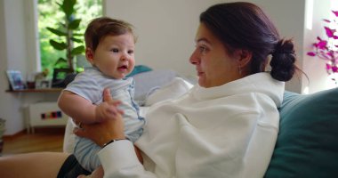 Bebeğin annesinin kollarındayken kameraya baktığı yakın plan, bebeğin parlak, ev ortamındaki meraklı ve nişanlı ifadesi..