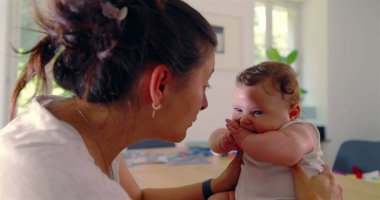 Anne bebeğini nazikçe kucağında tutuyor ve bir masada neşeli bir etkileşime giriyor. Bebek, aydınlık bir evde mutlu bir şekilde tepki veriyor.
