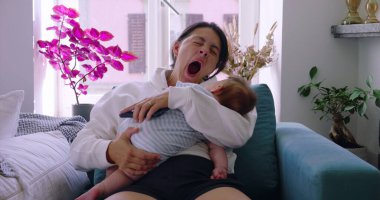 Anne, bebeğini koltukta emzirirken esniyor. Rahat bir ev ortamında, ebeveynlikte samimi bir yorgunluk ve sevgi anı yakalıyor.