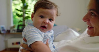 Bebeğin annesinin kollarındayken kameraya baktığı yakın plan, bebeğin parlak, ev ortamındaki meraklı ve nişanlı ifadesi..