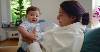 Bebeğin annesinin kollarındayken kameraya baktığı yakın plan, bebeğin parlak, ev ortamındaki meraklı ve nişanlı ifadesi..