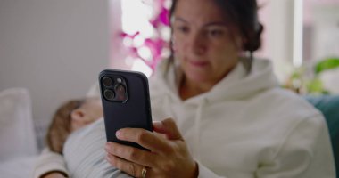 Yavrusu huzurlu bir oturma odasında, parlak doğal ışıkla kucağında dinlenirken bir annenin telefonunu kullanışının yakın çekimi..
