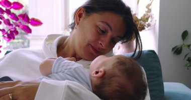 Anne bebeğini kollarında tutarken sıcak bir şekilde gülümsüyor. Yumuşak ışıkla sıcak bir ev ortamında şefkatli ve şefkatli bir an yaratıyor.