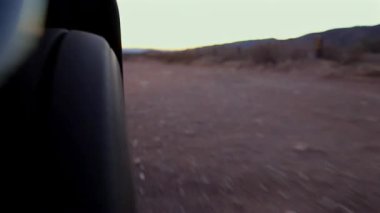 Gün batımında açık ve tozlu bir yolda hareket halindeki bir araç çarkı. Bulanık arka plan, engebeli arazide seyahat ederken hızı ve tekerleğe odaklanmayı vurgular.