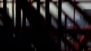 Şehir manzarası bulanık. Aşağı inen bir asansörün içindeki metal parmaklıklardan görülebiliyor. Endüstriyel yapı aşağıdaki hareketli şehirle çelişiyor..