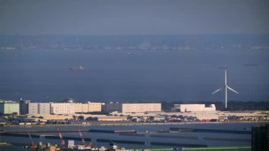 Uzakta rüzgar türbini ve gemilerinin bulunduğu kıyı sanayi bölgesi, sürdürülebilir enerji ve denizcilik operasyonlarının bir karışımını gösteriyor